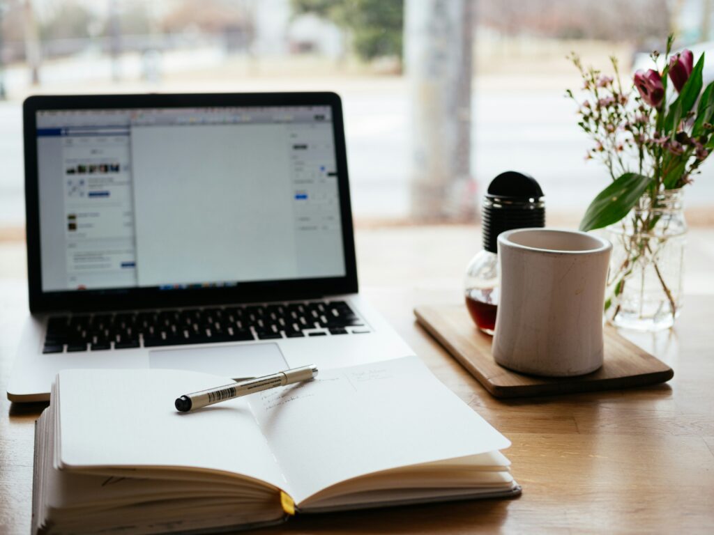 An image of a laptop and notebook lying on a wooden desk beside a cup of coffee and flower pot giving a soothing effect to the eyes of the viewer, representing the life style of a writer, through the segment behind the pages-Safar-e-adab